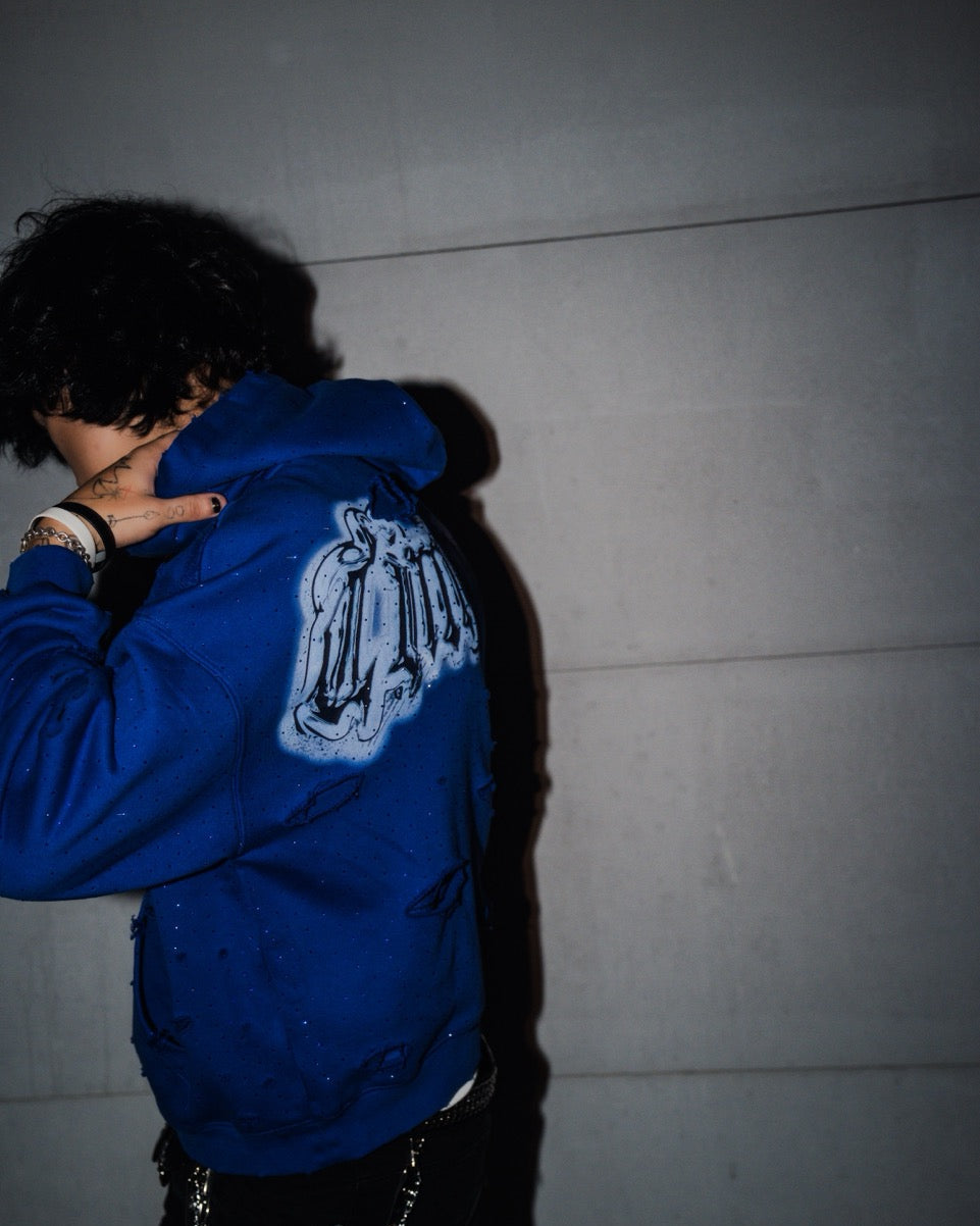 Person wearing a blue jacket with a logo on the back against a plain wall.