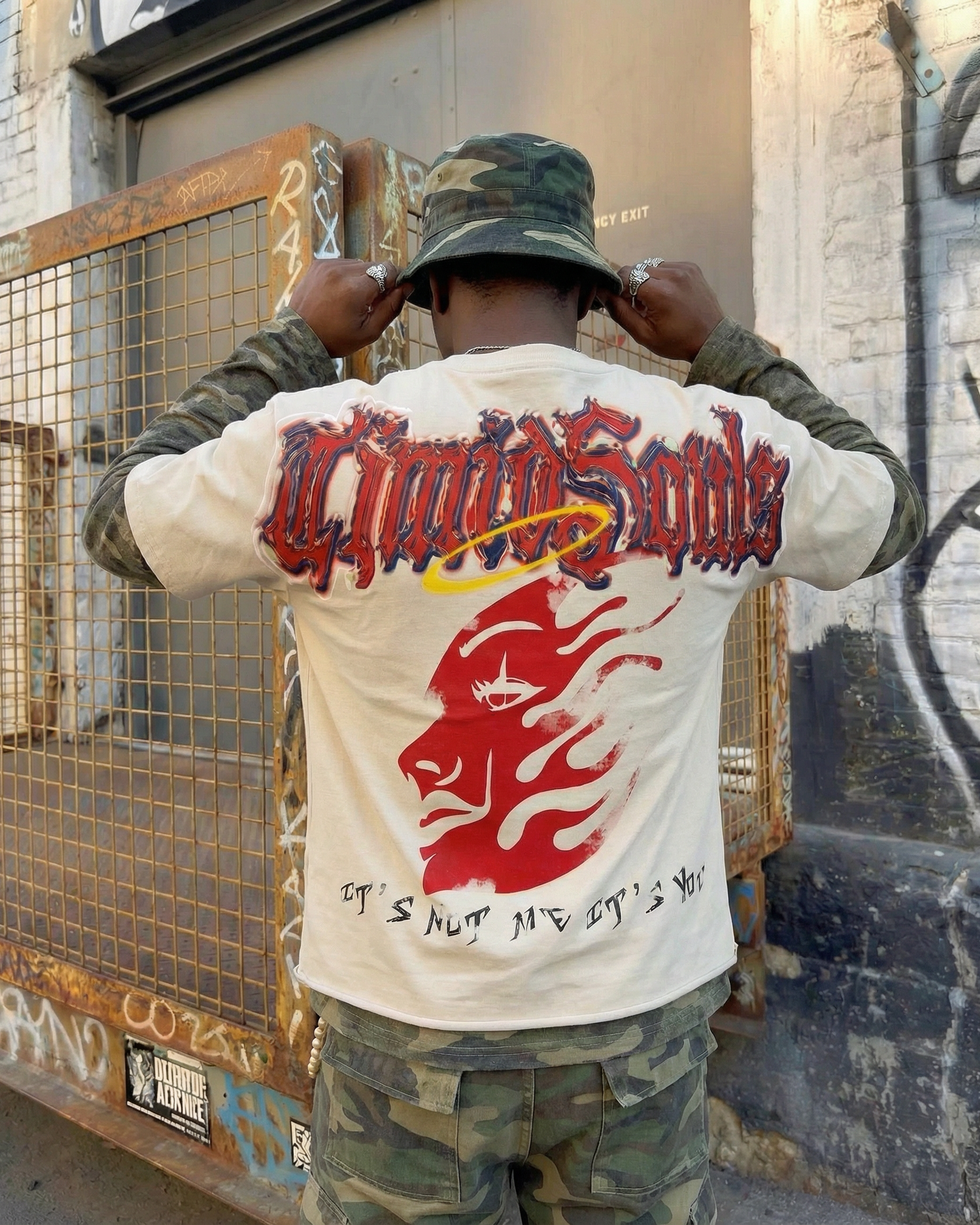 Person wearing a white long-sleeve shirt with red and black graphics and text, standing in front of a graffiti-covered wall.