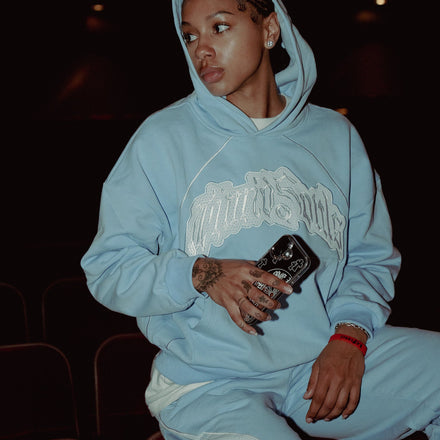 Person wearing a light blue hoodie with a logo, sitting in a dark room.