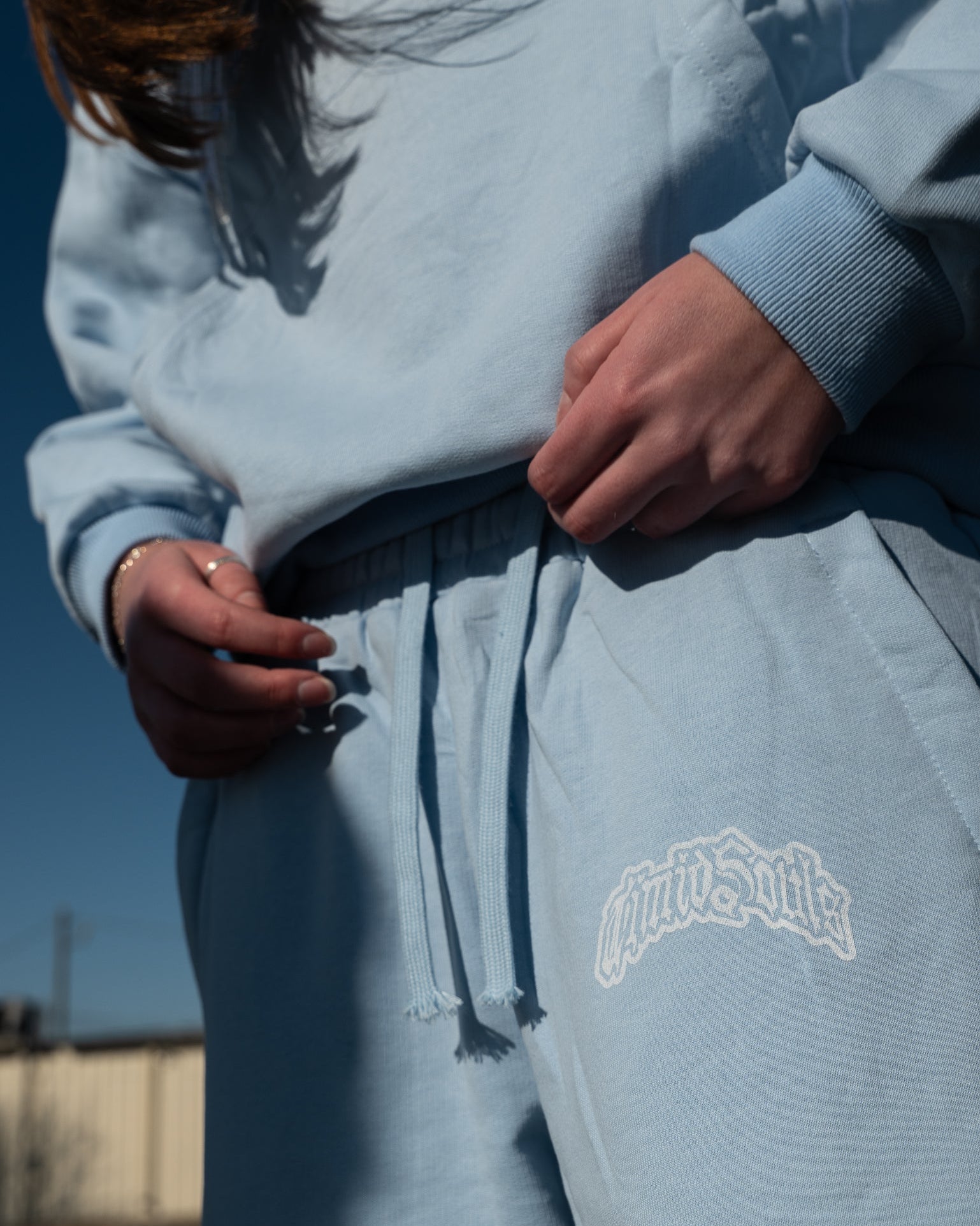Person wearing a light blue sweatshirt and pants with a brand logo, standing outdoors.