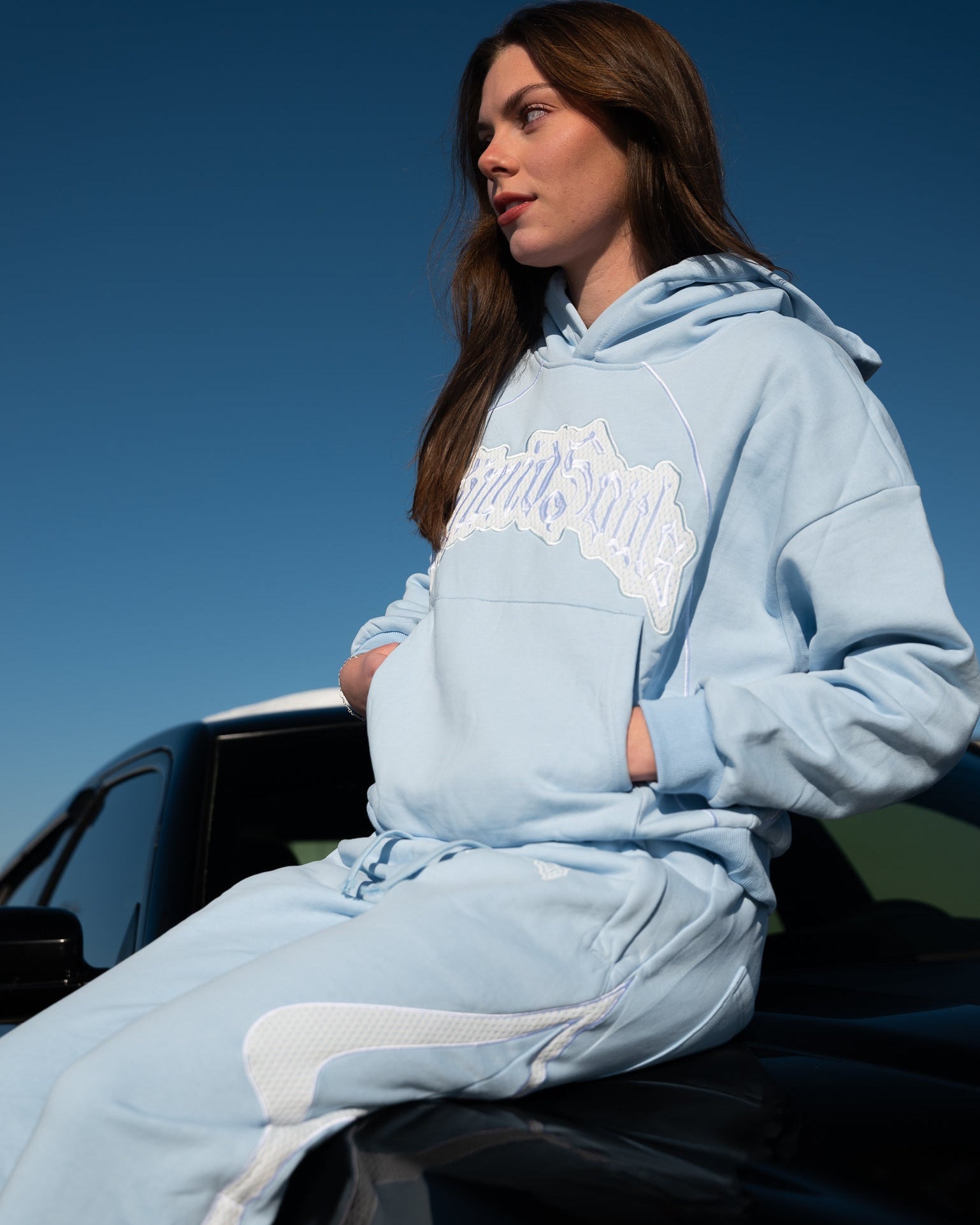 Person wearing a light blue hoodie with a logo, sitting on a car against a clear blue sky.
