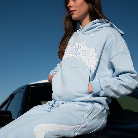 Person wearing a light blue hoodie with a logo, sitting on a car against a clear blue sky.