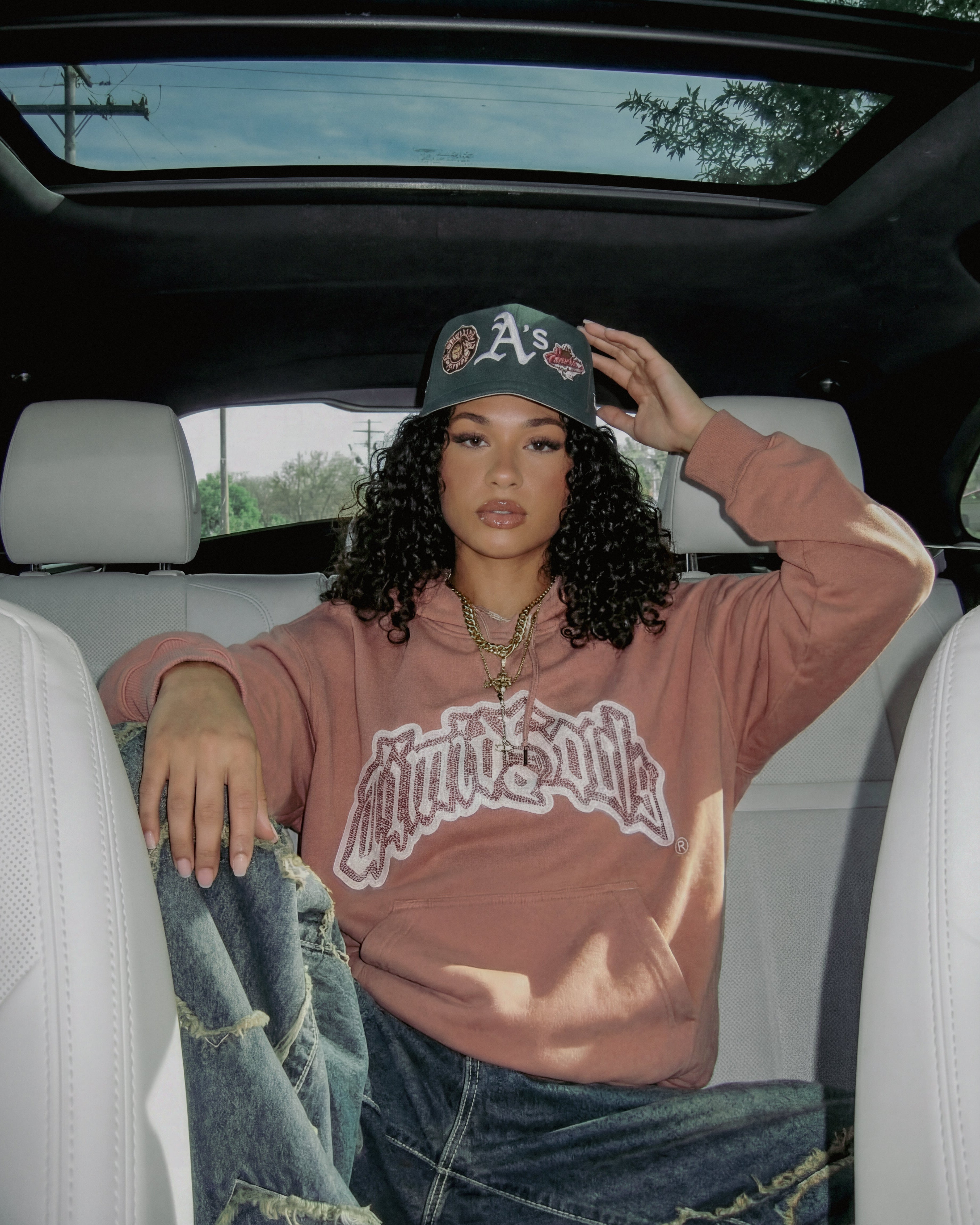 Person sitting in a car wearing a hoodie and cap, with a clear sky visible through the sunroof.