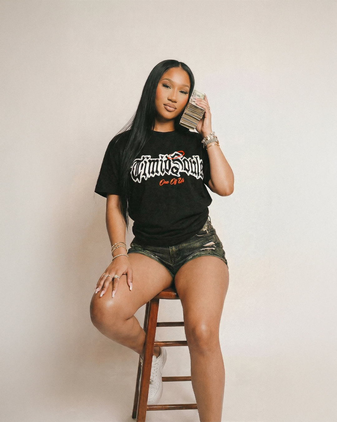 Woman wearing a black t-shirt with text, sitting on a wooden stool against a plain background