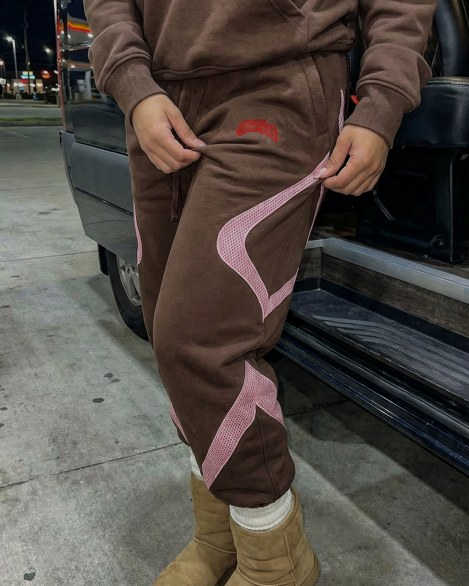 Person wearing a brown tracksuit with pink accents and tan boots standing next to a vehicle at night.