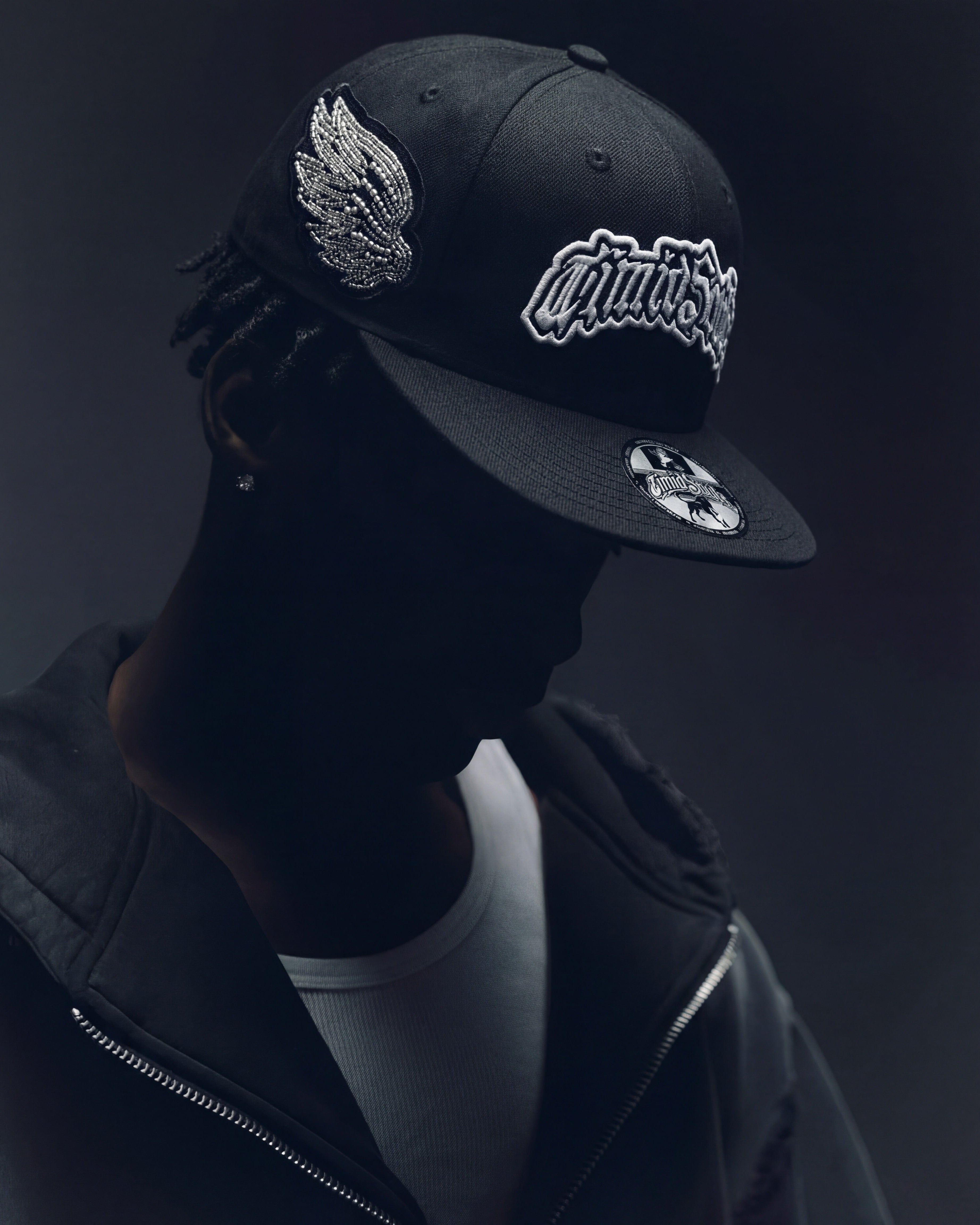 Black cap with wing emblem and brand logo worn by a person against a dark background