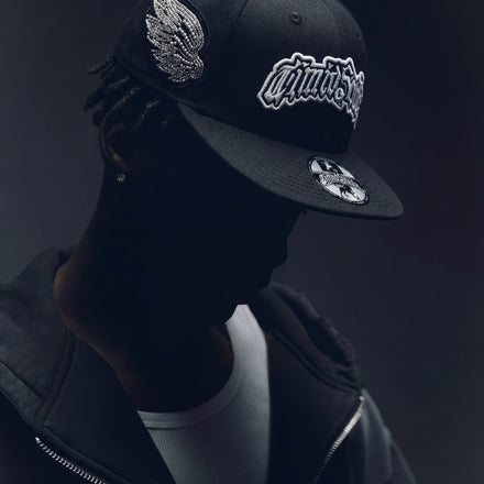 Black cap with wing emblem and brand logo worn by a person against a dark background